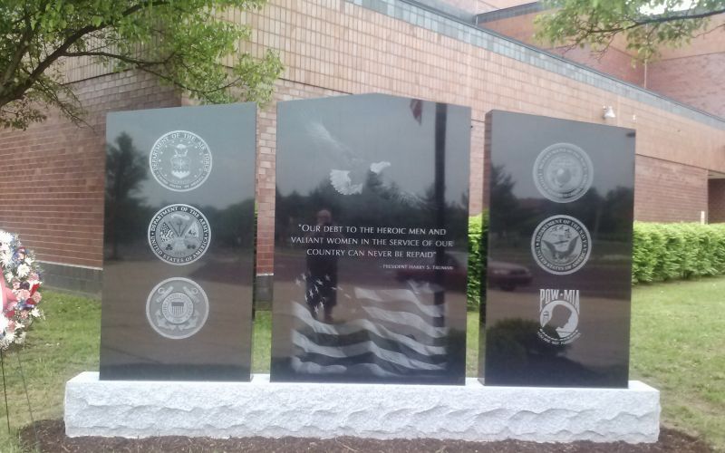 A three-paneled black granite war memorial resting on a rough-hewn, light gray stone base, situated in front of a brick building. Center Panel: Features etchings of a bald eagle in flight and a waving American flag, framing a quote from President Harry S. Truman: "Our debt to the heroic men and valiant women in the service of our country can never be repaid." Left Panel: Displays three vertically stacked seals for the Department of the Air Force, the Department of the Army, and the United States Coast Guard. Right Panel: Displays three vertically stacked emblems for the United States Marine Corps, the Department of the Navy, and POW-MIA.