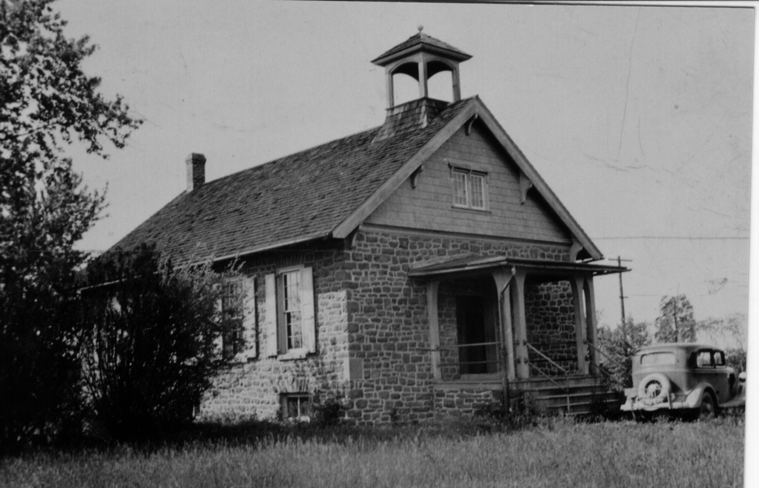 photo of one room school house with bell on top and old car parked to the right