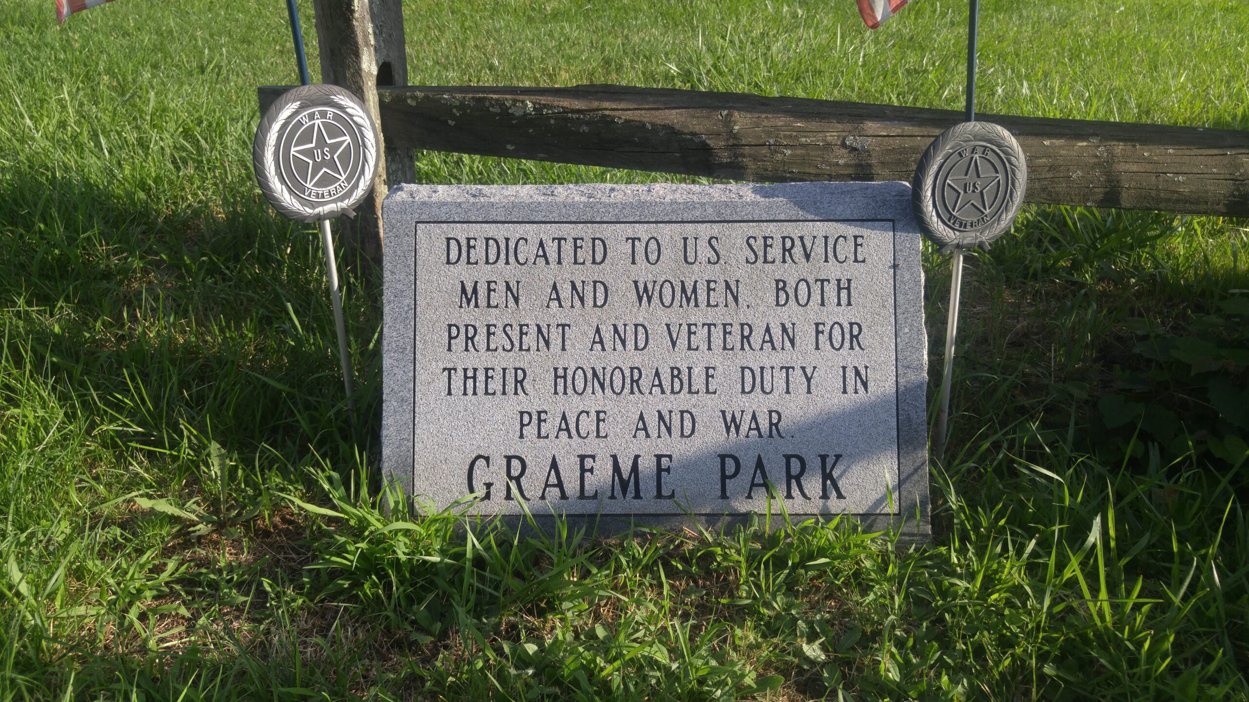 A light gray granite memorial stone resting in grass in front of a wooden split-rail fence, flanked by two circular metal "U.S. WAR VETERAN" markers. The inscription reads: "DEDICATED TO U.S. SERVICE MEN AND WOMEN, BOTH PRESENT AND VETERAN FOR THEIR HONORABLE DUTY IN PEACE AND WAR. GRAEME PARK"