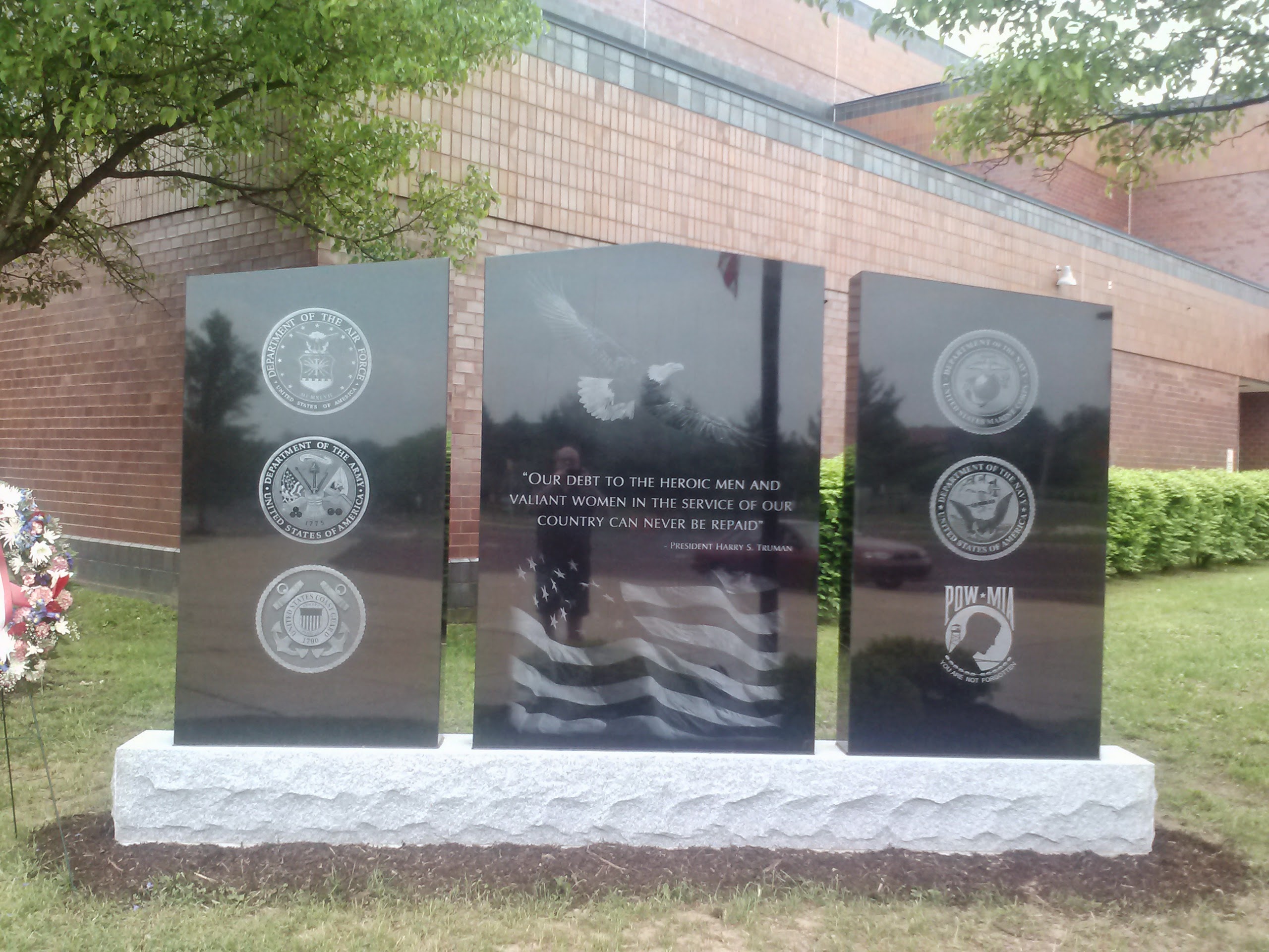 A three-paneled black granite war memorial resting on a rough-hewn, light gray stone base, situated in front of a brick building.  Center Panel: Features etchings of a bald eagle in flight and a waving American flag, framing a quote from President Harry S. Truman: "Our debt to the heroic men and valiant women in the service of our country can never be repaid."  Left Panel: Displays three vertically stacked seals for the Department of the Air Force, the Department of the Army, and the United States Coast Guard.  Right Panel: Displays three vertically stacked emblems for the United States Marine Corps, the Department of the Navy, and POW-MIA.