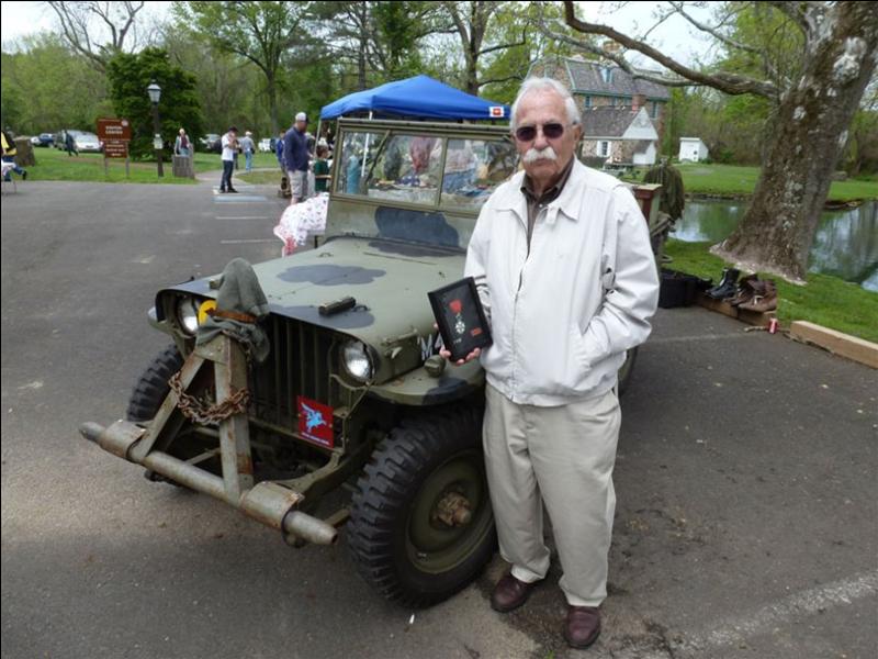 Herb Levy stands in the parking lot of Graeme Park next to a camouflage-painted 1942 Slat Grill Willys Jeep. He is wearing a white jacket and sunglasses, holding a framed military medal 'Cheveliar of the French legion of Honor'. In the background, the historic stone Keith Hoouse and a small pond are visible under a clear sky.
