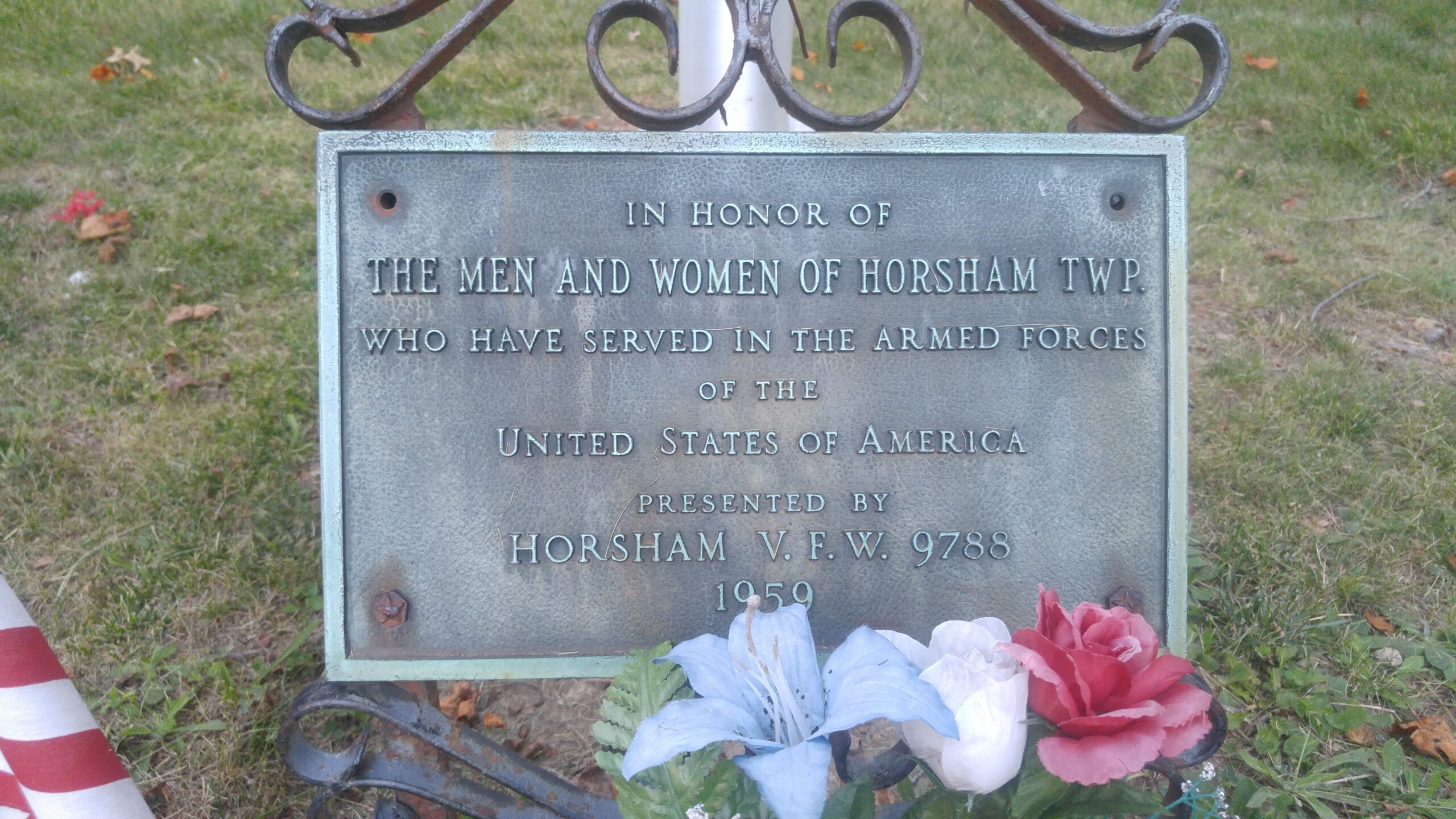 A weathered bronze memorial plaque mounted on a decorative metal stand outdoors, with artificial red, white, and blue flowers at its base. The plaque text reads: 