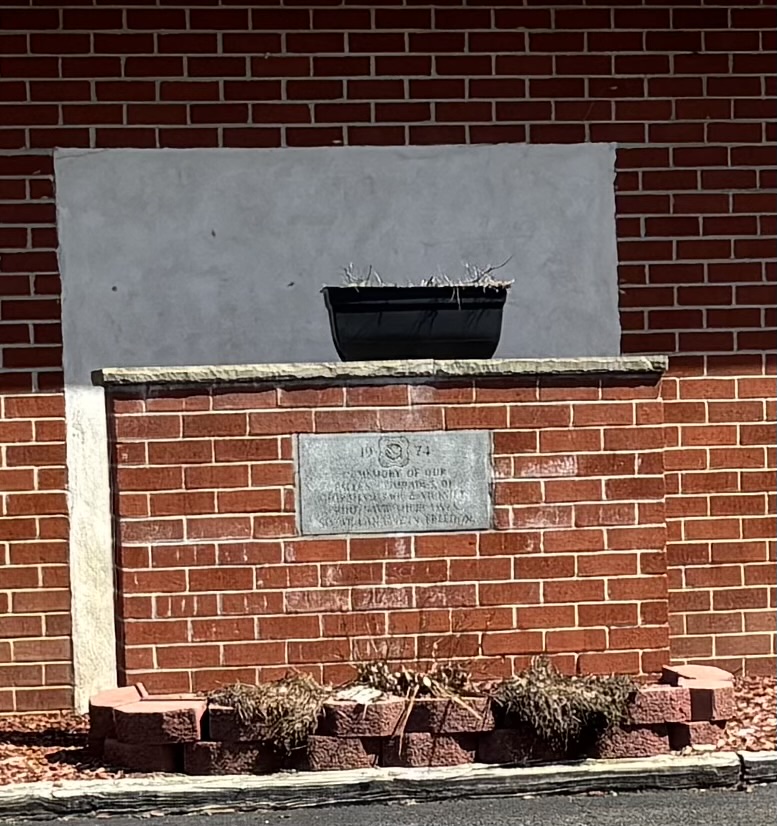 A weathered stone plaque embedded in a low brick monument, positioned in front of a brick building with a large rectangular patch of gray stucco. The plaque features a central seal flanked by the numbers 