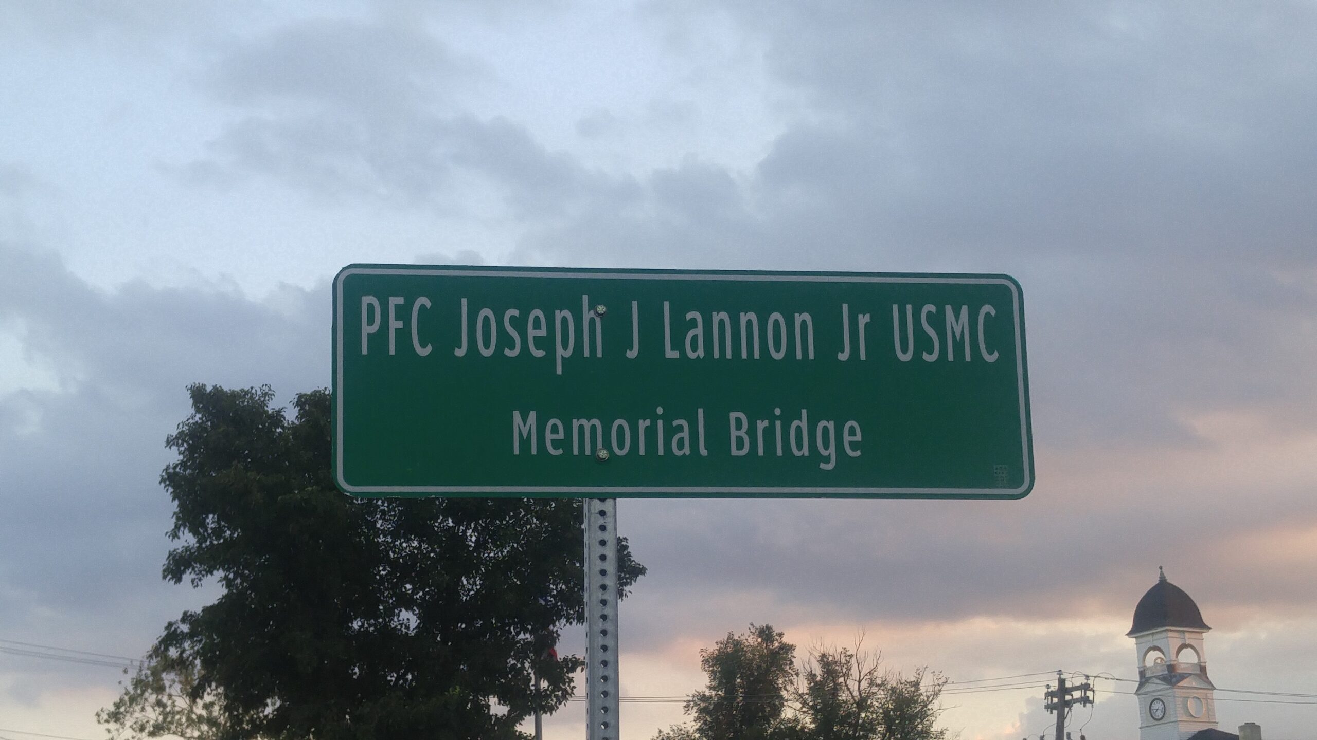 A green roadside memorial signnear the bridge at Horsham and York Rds that reads "PFC Joseph J Lannon Jr USMC Memorial Bridge." The sign is mounted on a metal post against a cloudy, twilight sky with trees and a white Loller Clock tower visible in the background.