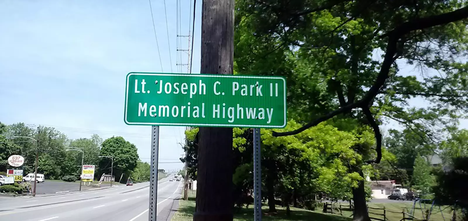 A green roadside sign reading "Lt. Joseph C. Park II Memorial Highway" mounted on a utility pole. The sign overlooks Easton Road (PA 611) with green trees and local business signs in the background.