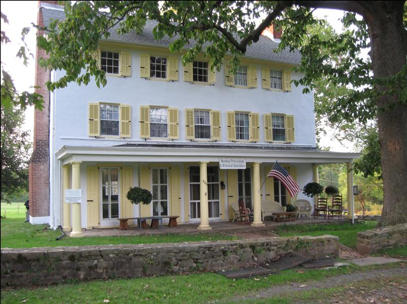 This photo shows the front exterior of the restored Penrose-Strawbridge House, a large, white three-story structure with yellow shutters on all windows. A long covered porch with columns runs across the first floor, furnished with several wicker chairs and a rocking chair. An American flag hangs from a pole on the porch. A small white sign above the porch entrance reads Horsham Preservation & Historical Association. Tree branches with green leaves hang down in the foreground and around the building, partially obscuring the upper floors and roof. The building is set against a backdrop of green trees.