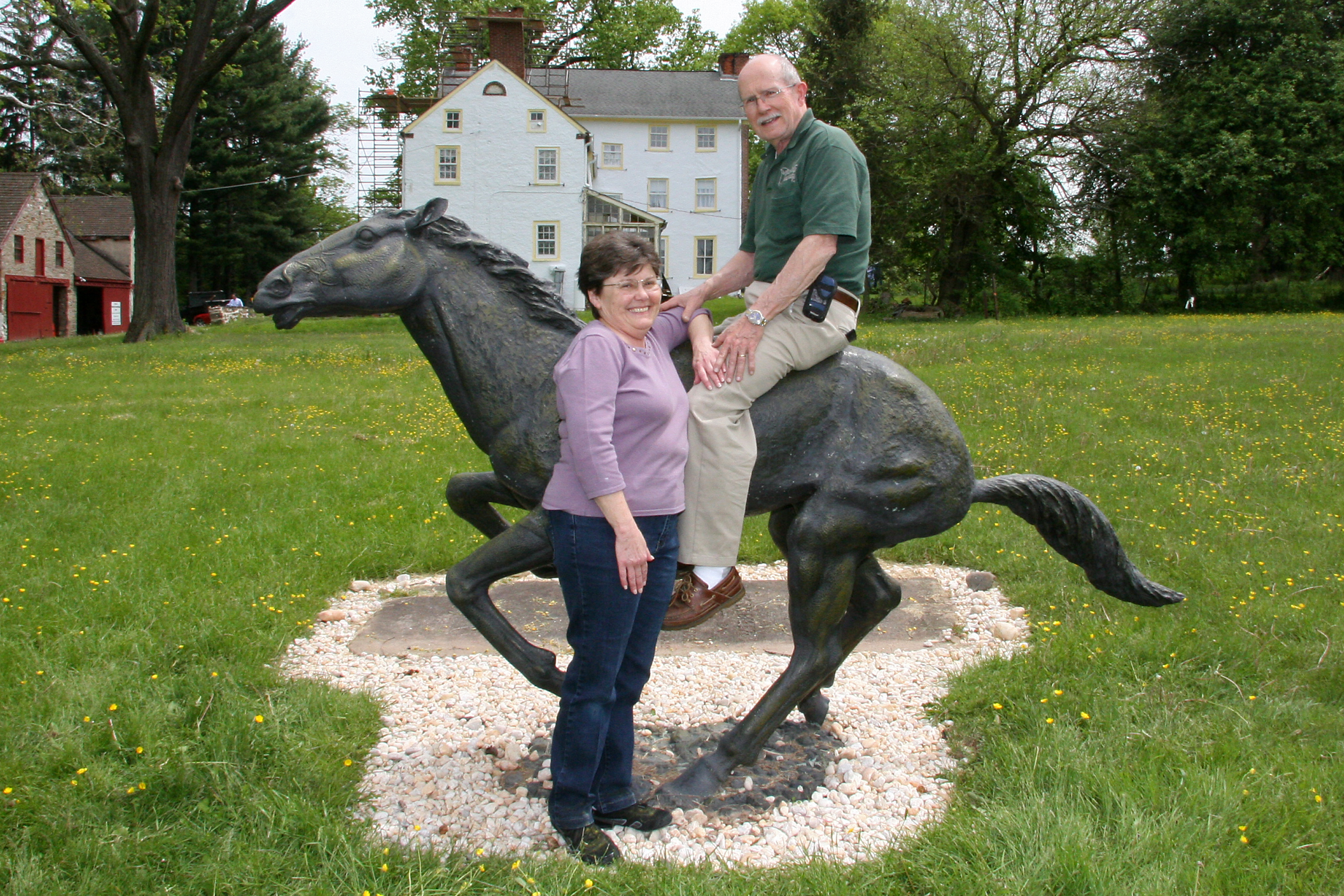 An outdoor photograph showing pete and margaret choate posing with a dark bronze horse statue in a grassy field. Margaret, with short brown hair and wearing a lavender long-sleeved shirt and dark blue jeans, stands smiling next to the horse, resting her hand on Pete's knee. Pete, wearing glasses, a green polo shirt, and khaki pants, sits on the horse's back, looking at the camera. The statue depicts a horse in mid-stride, with its front legs raised. They are situated on a patch of light-colored gravel. In the background, there is a large, white three-story stone building with yellow shutters (Penrose-Strawbridge House), along with a smaller red stone building and several large trees under an overcast sky.